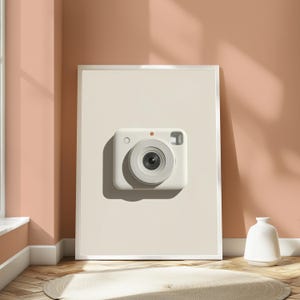 May include: A rectangular white framed wall art piece featuring a centered, minimalist illustration of a white instant camera. The camera has a prominent circular lens, a small orange dot above the lens, a circular button, and a rectangular viewfinder.