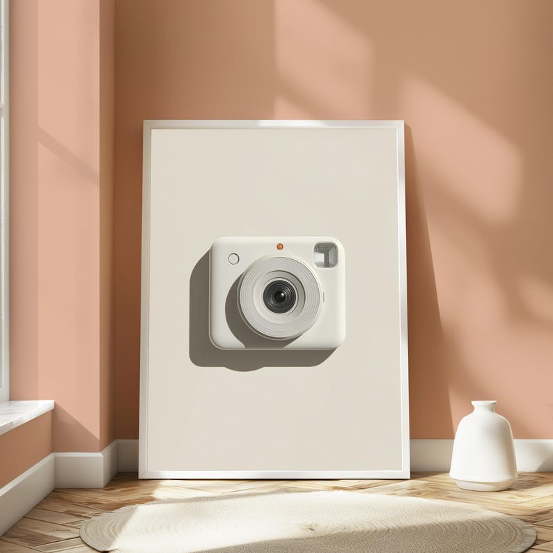 May include: A rectangular white framed wall art piece featuring a centered, minimalist illustration of a white instant camera. The camera has a prominent circular lens, a small orange dot above the lens, a circular button, and a rectangular viewfinder.