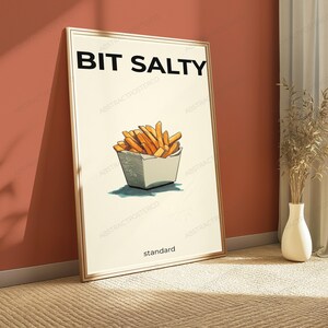 May include: A rectangular framed wall art print featuring a cream-colored background with the bold black text BIT SALTY at the top and the word standard at the bottom. In the center is a hand-drawn illustration of golden-brown french fries.