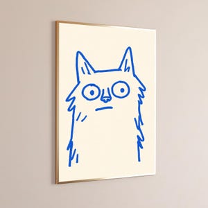 May include: A framed wall art print featuring a simple, hand-drawn illustration of a cat in vibrant blue ink on a cream-colored background. The cat has large, wide eyes, pointed ears, and a shaggy, textured outline, presented in a thin gold frame.