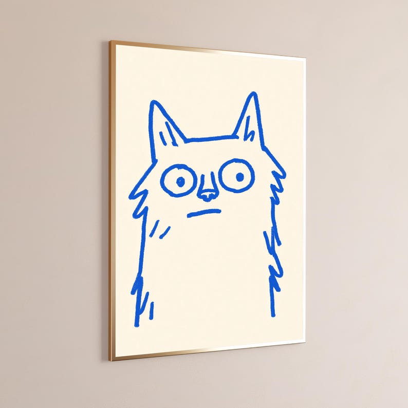 May include: A framed wall art print featuring a simple, hand-drawn illustration of a cat in vibrant blue ink on a cream-colored background. The cat has large, wide eyes, pointed ears, and a shaggy, textured outline, presented in a thin gold frame.