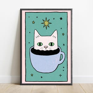May include: A framed art print featuring a light pink cat with green eyes sitting inside a light blue coffee cup. The background is a teal rectangle with a pink border, decorated with small black stars and a larger yellow star above the cat's head.
