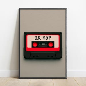 May include: A framed wall art print featuring a black and red cassette tape centred on a light grey background. The cassette tape has a white label with the text 2K POP written in black ink and two red circular spools visible in the centre.