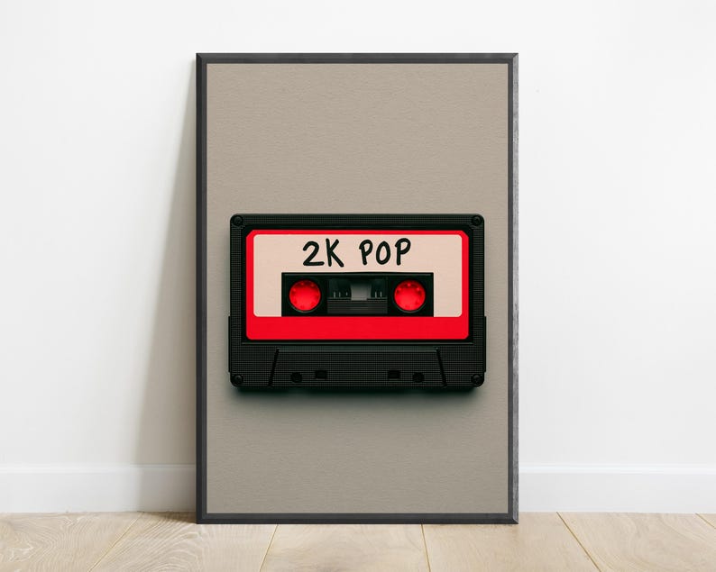 May include: A framed wall art print featuring a black and red cassette tape centred on a light grey background. The cassette tape has a white label with the text 2K POP written in black ink and two red circular spools visible in the centre.