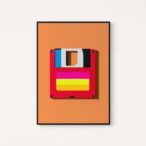 May include: A framed wall art print featuring a red floppy disk centred on a solid orange background. The disk has a white sliding metal shutter, a blue and black top section, and a label with horizontal pink, yellow, and orange stripes.