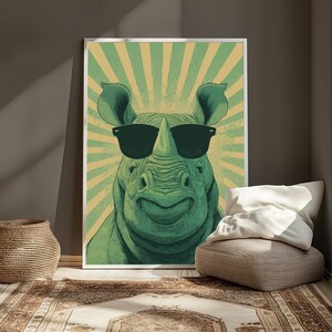 May include: A framed art print featuring a green rhinoceros wearing black sunglasses. The rhino is smiling against a yellow and green sunburst background. The artwork is displayed on a wall, leaning against a neutral-toned wall.