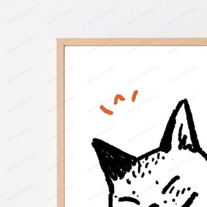 Pu&ograve; includere: Una stampa artistica da parete incorniciata raffigurante un disegno a inchiostro nero di una testa di gatto con linee strutturate e abbozzate. Sopra il gatto, tre piccoli segni a trattino curvo di colore arancione. Cornice in legno chiaro.