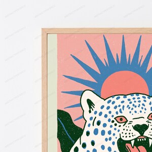 May include: A framed wall art print featuring a white leopard with blue spots, a dark green leaf, and a pink sun with blue rays against a light pink background. The print is enclosed in a light-colored wooden frame with a thin vertical light green border.