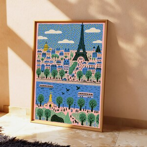 K&ouml;nnte beinhalten: Ein gerahmter Kunstdruck mit einer stilisierten Illustration der Pariser Skyline, der Eiffelturm, einem blauen Fluss, gr&uuml;nen B&auml;umen und Geb&auml;uden. Die Szene zeigt kleine Figuren, Busse und wei&szlig;e Wolken vor einem gelb gepunkteten blauen Himmel.