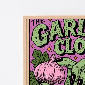 Pu&ograve; includere: Una stampa artistica da parete incorniciata raffigurante un bulbo d'aglio rosa e una mano verde con lunghe unghie nere su sfondo viola. Il testo recita THE GARLIC CLOVE in grandi lettere verdi con contorni neri, accentuato da piccole stelle arancioni.