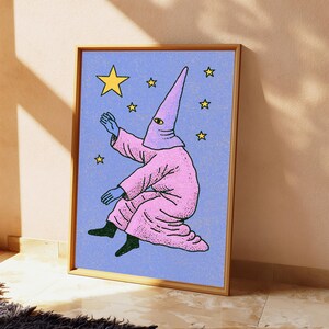 May include: A framed art print featuring a figure in a pink hooded robe with a single visible eye, reaching toward yellow stars against a textured light blue background. The figure has blue hands and dark feet, rendered in a graphic illustration style.