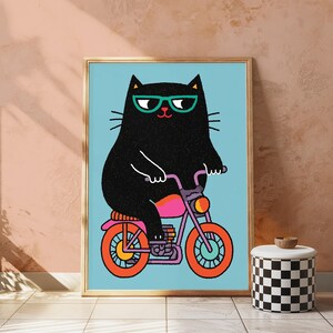 K&ouml;nnte beinhalten: Ein gerahmter Kunstdruck, der eine schwarze Katze mit t&uuml;rkisfarbener Brille auf einem violetten Motorrad mit orangefarbenen R&auml;dern vor hellblauem Hintergrund zeigt. Die Illustration verwendet kr&auml;ftige Linien und leuchtende Farben.