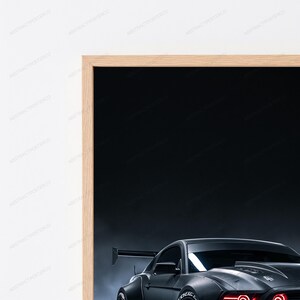 May include: A framed wall art print featuring a dark grey sports car with a prominent rear spoiler and glowing red headlights against a dark, smoky background. The artwork is enclosed in a light-toned wooden frame, showcasing a sleek automotive design.