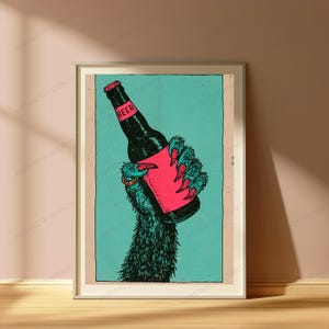 May include: A framed art print featuring a dark, furry creature hand with sharp claws holding a black beer bottle. The bottle has a pink label with the word BEER printed in black. The background is a solid teal colour within a rectangular border.