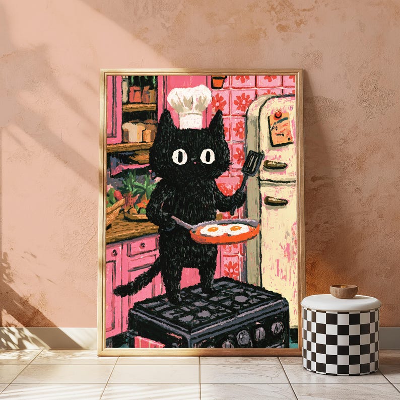 Peut inclure: Une impression d'art encadr&eacute;e repr&eacute;sentant un chat noir portant une toque de chef blanche, debout sur une cuisini&egrave;re noire tout en tenant une po&ecirc;le avec deux &oelig;ufs au plat. Le fond montre une cuisine rose avec des placards et des carreaux.
