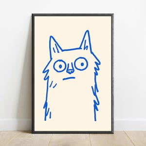 May include: A framed wall art print featuring a simple, bold blue line drawing of a cat with wide, circular eyes and a straight mouth. The illustration is centered on a light cream-colored background, enclosed within a thin black frame.