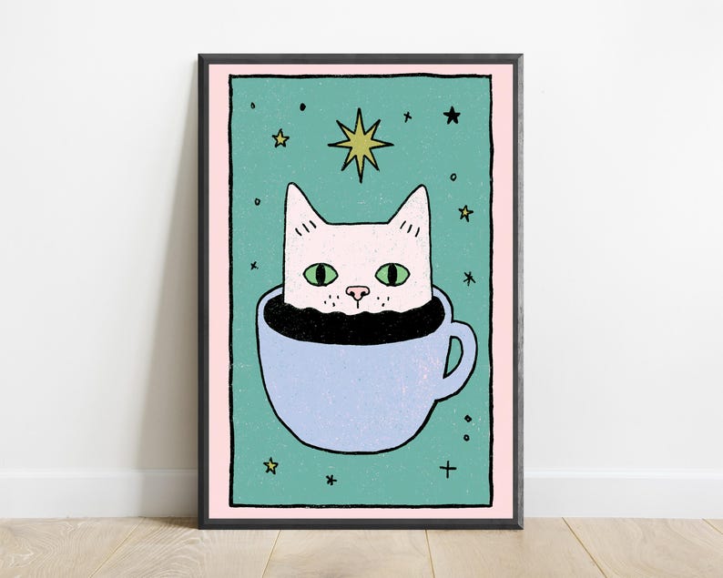 May include: A framed art print featuring a light pink cat with green eyes sitting inside a light blue coffee cup. The background is a teal rectangle with a pink border, decorated with small black stars and a larger yellow star above the cat's head.