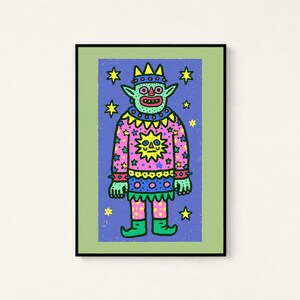 May include: A framed art print featuring a green creature with a crown, wearing a pink sweater with a yellow sun and star pattern. The figure stands against a blue background filled with yellow stars, all enclosed in a light green border.