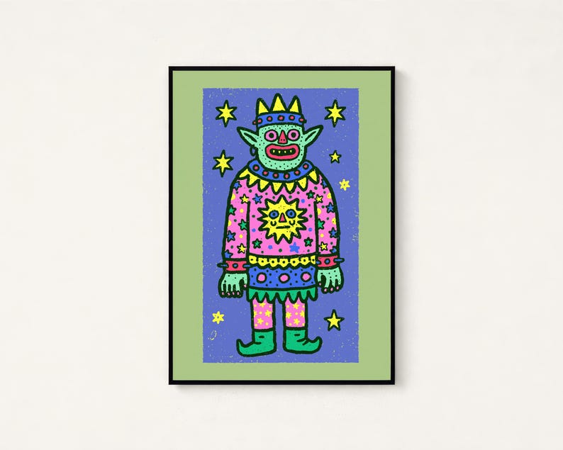 May include: A framed art print featuring a green creature with a crown, wearing a pink sweater with a yellow sun and star pattern. The figure stands against a blue background filled with yellow stars, all enclosed in a light green border.