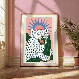 May include: A framed art print featuring a white leopard with blue spots, shown with its mouth open, set against a pink background with a blue sunburst graphic and dark green leaves. The leopard has pink eyes and a pink tongue, framed in gold.