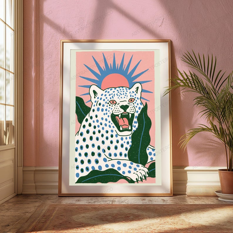 May include: A framed art print featuring a white leopard with blue spots, shown with its mouth open, set against a pink background with a blue sunburst graphic and dark green leaves. The leopard has pink eyes and a pink tongue, framed in gold.