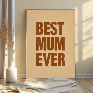 May include: A framed print featuring the text "BEST MUM EVER" in brown, bold letters against a beige backdrop. The image is set in a room with a vase of dried plants and a white cloth.