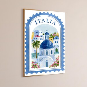 May include: A framed wall art print featuring a watercolor illustration of a Mediterranean-style building with a blue dome, a palm tree, and pink flowers, framed by a blue postage stamp border with the word ITALIA printed at the top.