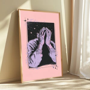 May include: A framed rectangular wall art print featuring a light pink background and a central black rectangular panel. The illustration depicts two hands held up to a face, rendered in light purple with black line work, set against a starry night sky.