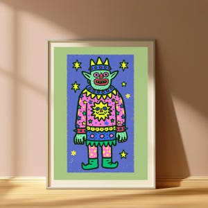 May include: A framed art print featuring a green creature with pointed ears wearing a crown and a pink sweater decorated with a yellow sun and stars. The creature has a blue skirt and green boots, set against a blue background with yellow stars.