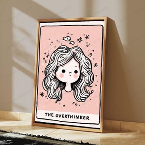 May include: A framed wall art print featuring a light pink background with a black line drawing of a person with wavy hair. Above the head are small doodles of fish and stars. The text THE OVERTHINKER is printed in black at the bottom of the print.
