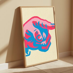 May include: A framed wall art print featuring a stylised, abstract illustration of a bull in vibrant shades of hot pink, light pink, and bright blue against a cream-coloured background. The artwork is encased in a simple, light-toned wooden frame.
