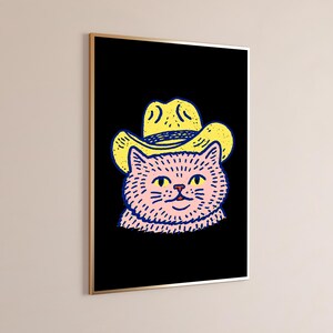 Puede incluir: Una impresi&oacute;n de arte mural enmarcada que presenta una ilustraci&oacute;n gr&aacute;fica de un gato rosa con un sombrero de vaquero amarillo sobre un fondo negro s&oacute;lido. El gato tiene ojos amarillos y contornos azul oscuro, en un marco met&aacute;lico fino.