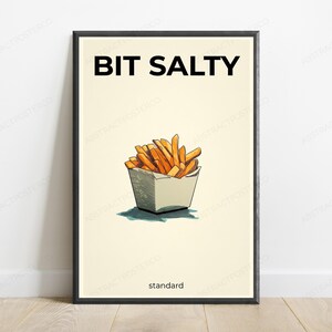 May include: A framed wall art print featuring a cream background with a black border. The text BIT SALTY appears in black at the top, and the word standard is at the bottom. A central illustration shows golden-brown french fries in a white container.