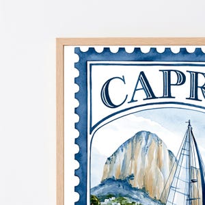 May include: A framed watercolor print designed as a postage stamp featuring the text CAPRI. The artwork depicts a sailboat with white sails positioned in front of a large, tan-colored rocky mountain under a light blue sky with white clouds.