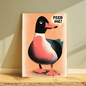 May include: A framed art print featuring a stylised illustration of a duck with a black head, white neck, and pinkish-orange chest and feet. The duck has a dark teal wing and a speech bubble above its head containing the black text FEED ME!