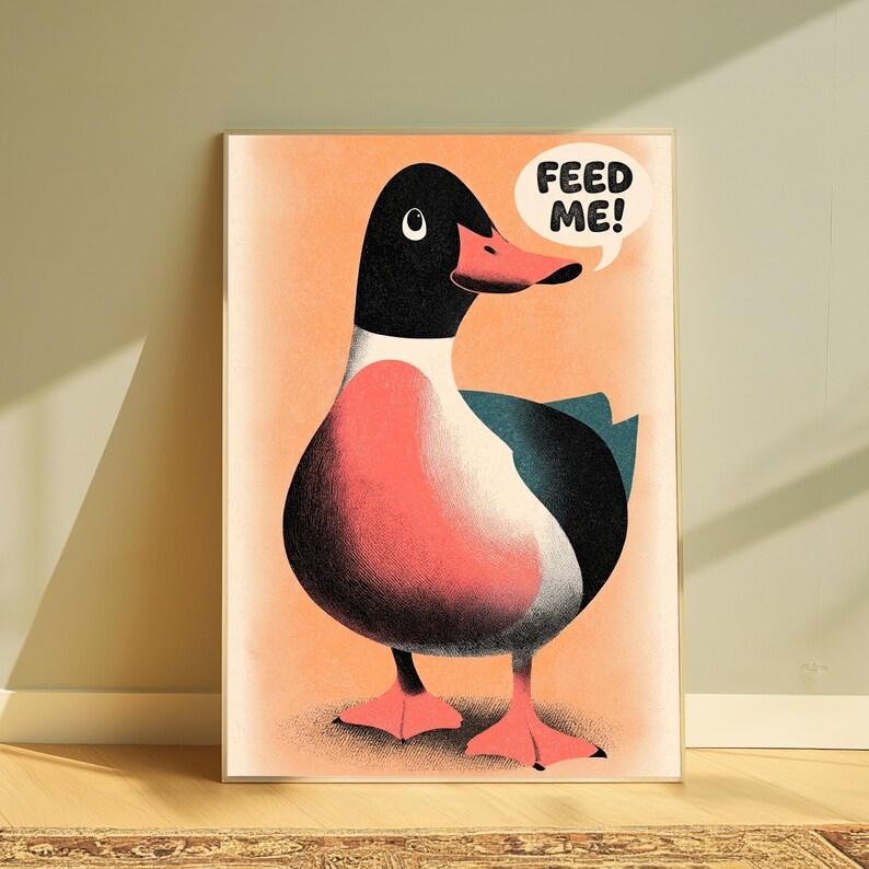 May include: A framed art print featuring a stylised illustration of a duck with a black head, white neck, and pinkish-orange chest and feet. The duck has a dark teal wing and a speech bubble above its head containing the black text FEED ME!
