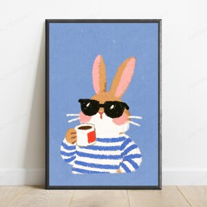 Op de afbeelding: Een ingelijste wandkunstprint met een lichtbruin konijn met roze oren, een zwarte zonnebril en een blauw-wit gestreept shirt met lange mouwen. Het konijn houdt een kleine witte mok met een rood vierkant detail vast tegen een effen blauwe achtergrond.