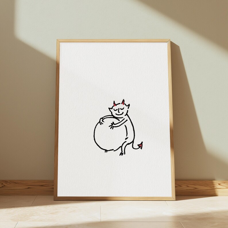 May include: A framed wall art print featuring a simple black line drawing of a small devil character with red horns and a red tail tip, hugging a large circular shape. The illustration is centred on a plain white background within a light wood frame.