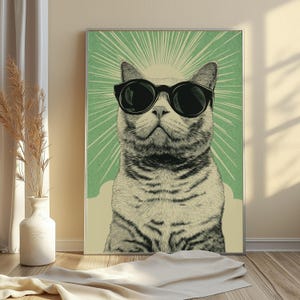 May include: A framed wall art print featuring a black and white illustration of a cat wearing large black sunglasses. The cat is centered against a light green background with radiating white sunburst lines and a solid cream-colored base section.