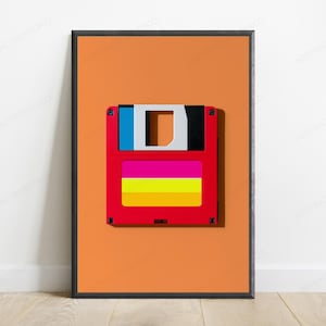 May include: A framed wall art print featuring a centered, stylized floppy disk graphic against a solid orange background. The disk is red with a blue and black top section, and a central label area displaying horizontal pink, yellow, and orange stripes.
