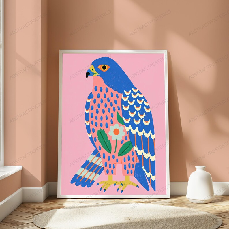 May include: A framed wall art print featuring a stylized blue and pink falcon illustration. The bird has a bright yellow eye, a black beak, and yellow talons. A single white flower with green leaves is centered on the bird's chest against a pink background.