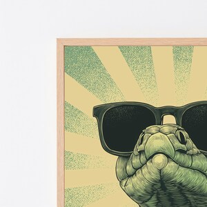 May include: A framed graphic art print featuring a green snake wearing large black sunglasses. The background consists of a pale yellow sunburst pattern with a stippled texture. The frame is made of light-colored wood with a visible grain.