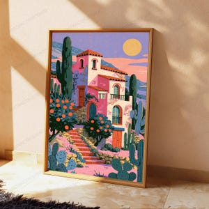 May include: A framed art print featuring a pink Mediterranean-style villa with arched windows and doors, surrounded by orange trees and tall green cacti. The scene includes a stone staircase leading to the house under a pale yellow sun and purple sky.