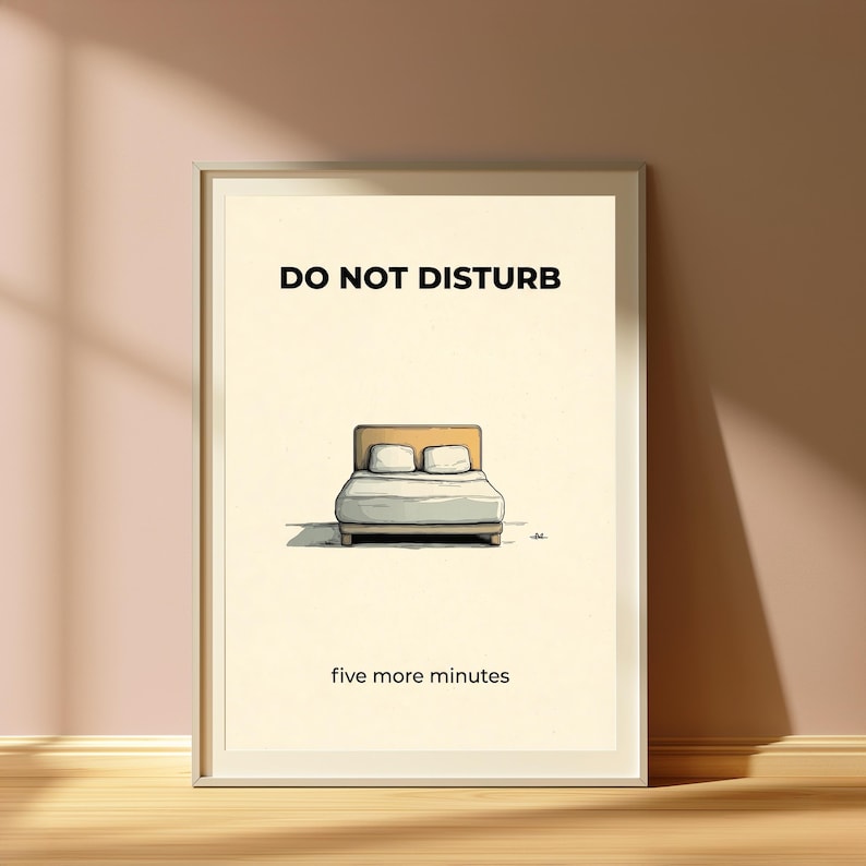 Puede incluir: Un cuadro enmarcado con las palabras "DO NOT DISTURB" sobre una ilustraci&oacute;n de una cama con dos almohadas. Debajo de la ilustraci&oacute;n de la cama, el texto dice "five more minutes". La obra de arte est&aacute; sobre un fondo crema.