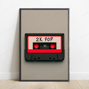 Könnte beinhalten: Ein gerahmter Wandkunstdruck mit einer schwarzen Kassette, einem roten Etikett in der Mitte und dem in schwarzer Tinte geschriebenen Text 2K POP. Die Kassette ist auf einem einfarbigen, hellbeigen Hintergrund zentriert und von einem dünnen schwarzen Rahmen umgeben.
