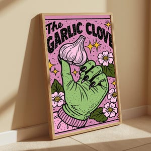 May include: A framed art print featuring a green hand with black fingernails holding a pink garlic clove against a pink background with flowers and yellow stars. The text at the top reads The GARLIC CLOVE in bold black letters with a dotted pattern.