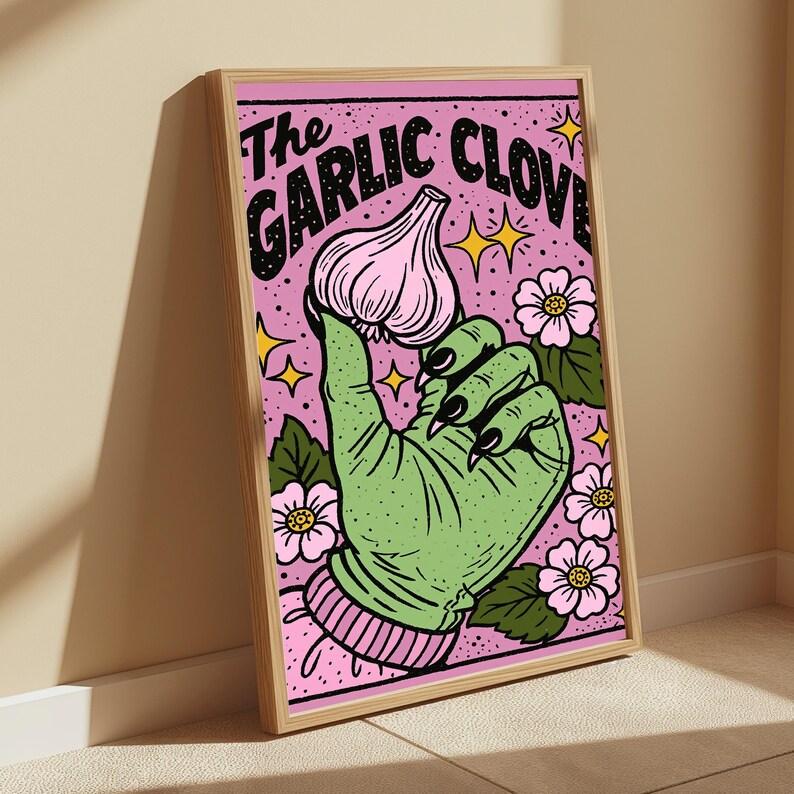 May include: A framed art print featuring a green hand with black fingernails holding a pink garlic clove against a pink background with flowers and yellow stars. The text at the top reads The GARLIC CLOVE in bold black letters with a dotted pattern.