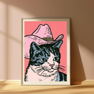 May include: A framed art print featuring a graphic illustration of a black and white tabby cat wearing a light pink cowboy hat. The cat has a white chest and face markings, set against a solid coral pink background with bold black outlines.