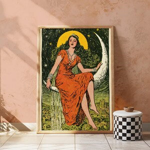 May include: A framed art print featuring a person with long dark hair wearing a rust-orange dress, seated on a large crescent moon against a dark green starry sky with a yellow circular halo behind their head and a landscape visible below.