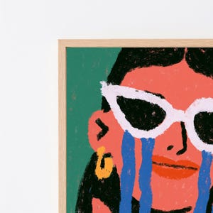 May include: A framed art print featuring a stylized illustration of a face with dark hair, wearing white cat-eye sunglasses. The figure has coral-colored skin, a bright orange earring, and thick blue streaks resembling tears on the cheeks.